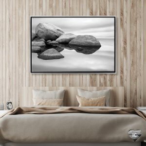 Framed black and white photo of of large granite stones above water by iCanvas artist Jeff Friesen mounted above a bed